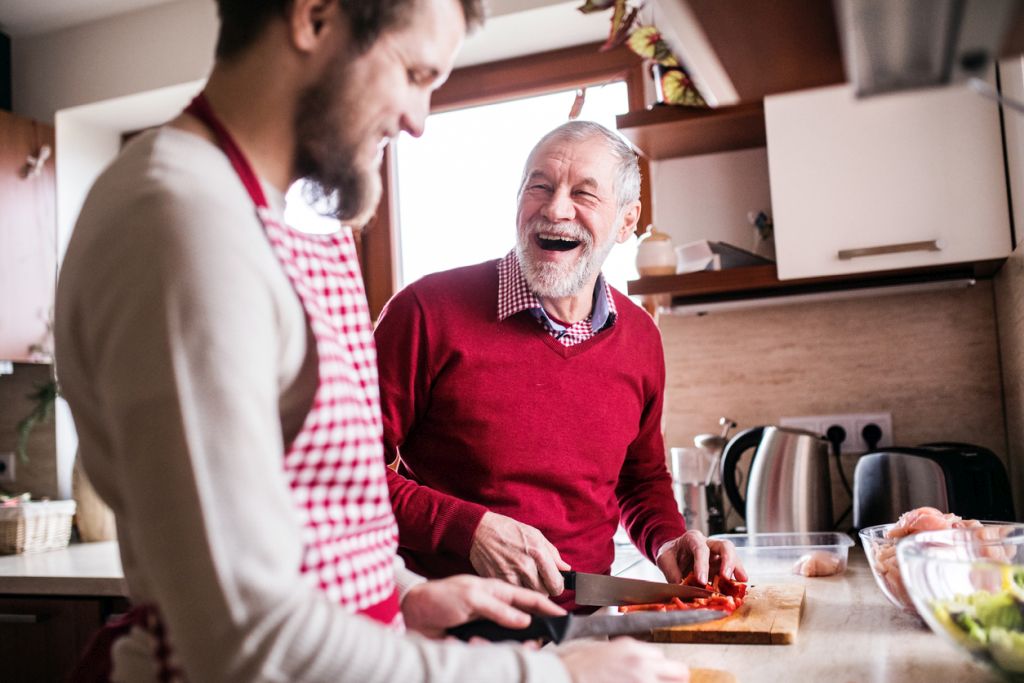 Illustrative image of a person laughing and chopping a pepper while interacting with another person in a home kitchen.