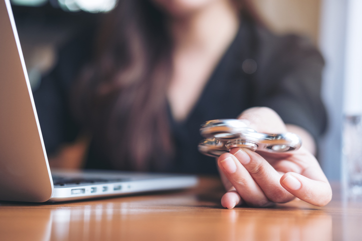 Illustrative image of a person using a laptop while playing with a fidget spinner on a wooden table.