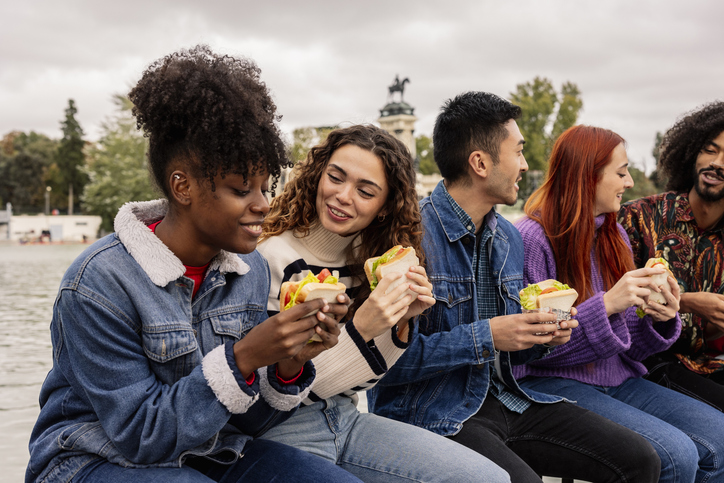 Illustrative image of a group of people eating sandwiches together outside.