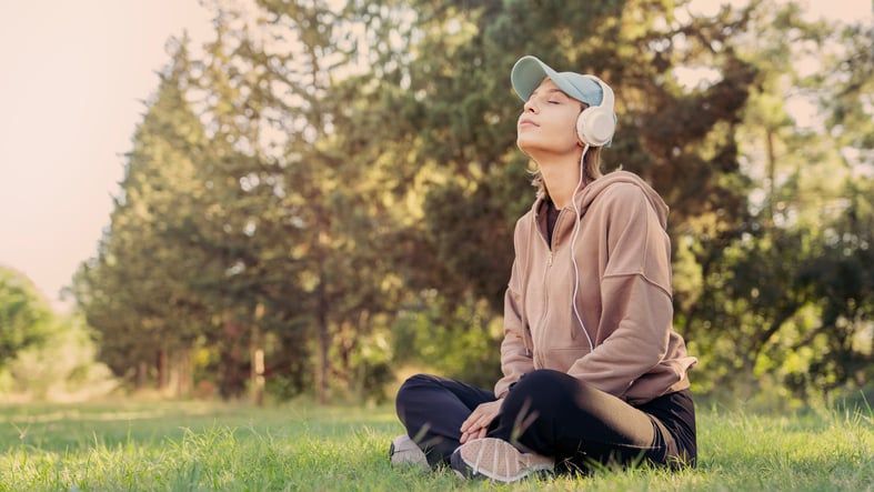 Illustrative photo of a person sitting on the grass in the lotus position, eyes closed, and listening to their headphones.