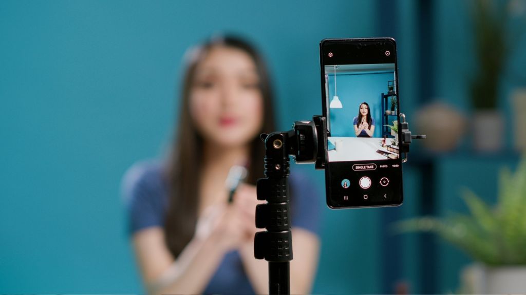 Illustrative image of a person standing in front of a phone on a tripod, with their blurred image in the background and their clear image displayed on the phone screen in the foreground.