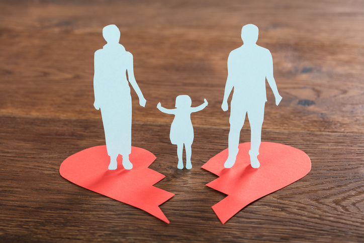 Illustrative image of paper cut outs of a child in between two separated adult figures, with each adult figure standing on one half of a broken paper heart.