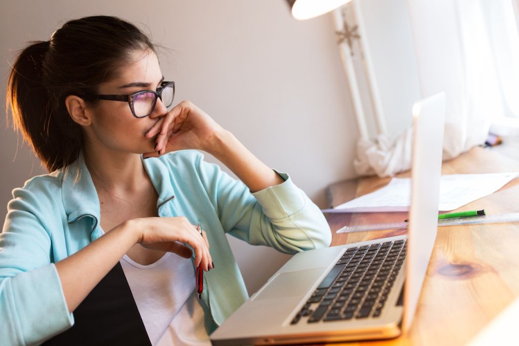 Illustrative image of a person seated at a table, looking at their laptop with a pen in one hand and the other hand held to their mouth, showing a pensive expression.