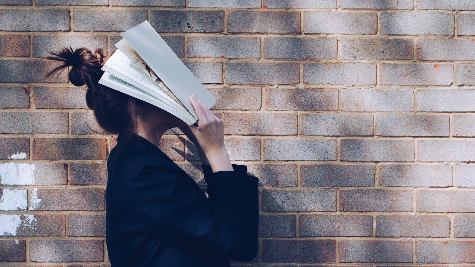 Illustrative image of a person standing next to a brick wall with a book over their head.