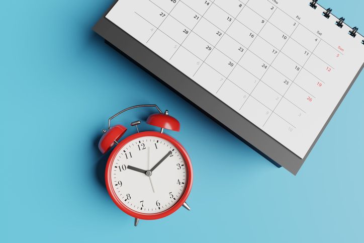 Illustrative image of a classic red alarm clock next to a paper calendar on a desk.