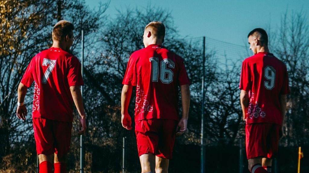Illustrative image of the backs of three people walking together on a football pitch, wearing red jerseys numbered 7, 16, and 9 from left to right.