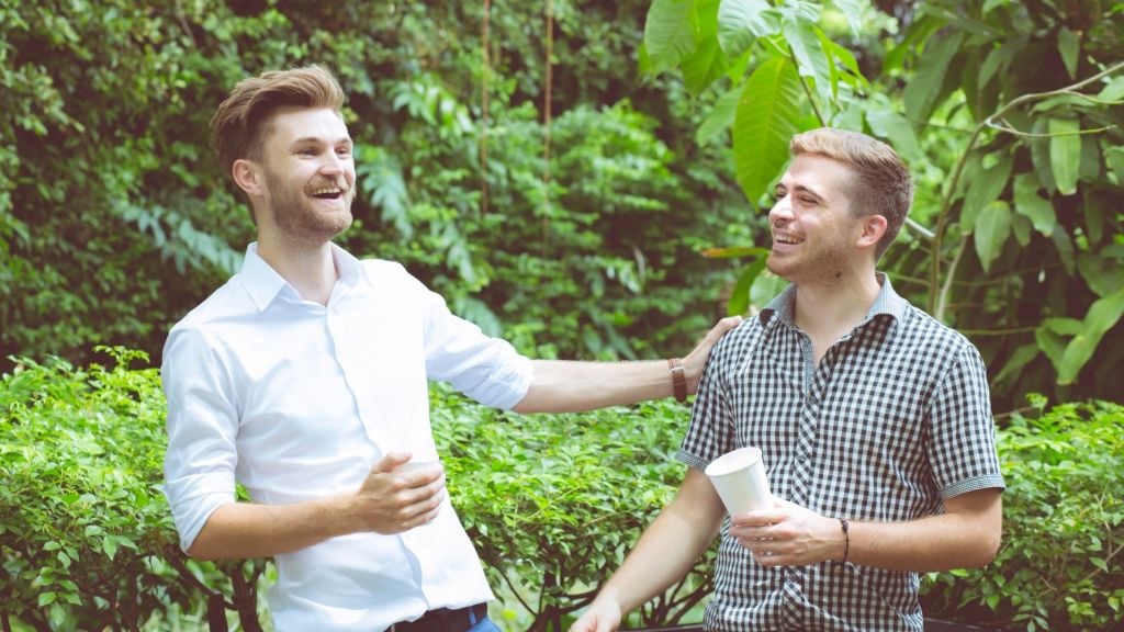Illustrative image of two smiling people talking outdoors in a green area, with one person clapping the other's shoulder.