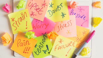 Different coloured sticky notes stuck to a notepad, featuring the words "upset," "doubt," "pressure." "stress," "worry," "strain," "pain," "tension" and "failure."