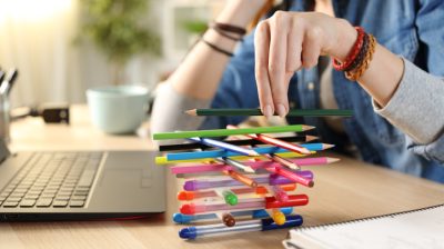 Illustrative image of a person building a pencil tower while seated in front of their laptop.
