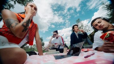 a group of students sit on a picnic blanket, one is listening to music, another talks on the phone