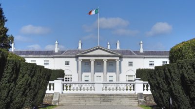 White building with columns out front and an Irish flag hanging overhead, Áras an Uachtaráin the official residence and principal workplace of the President of Ireland.