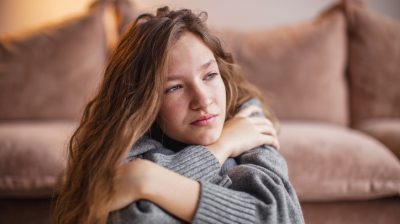 Illustrative image of a person crouching in front of a couch with their arms wrapped around their chest.