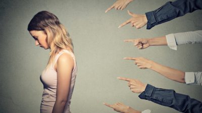 Illustrative image of 6 different hands pointing towards a sad person with back turned and head bowed.