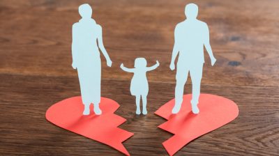 Illustrative image of paper cut outs of a child in between two separated adult figures, with each adult figure standing on one half of a broken paper heart.