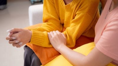 Illustrative close-up image of a person comforting a friend on a sofa in a living room.