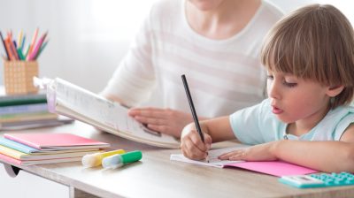 Illustrative image of a child focusing on homework with an adult in the background.