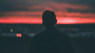 A silhouette of a person standing at the edge of a calm lake during sunset, with warm golden and orange hues in the sky and water, evoking a sense of solitude and reflection.