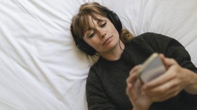 Illustrative image of a person lying on a bed while viewing their phone and listening to music through headphones.