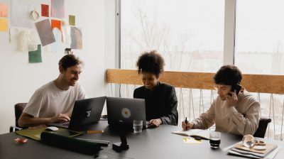 three people sit in a bright office space at a table with laptops and notebooks co-working