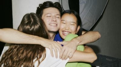 three people pose for a ground shot two face the camera and one face away. They are hugged and the central person and person to the right have huge smiles.