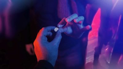 Hand of a person being handed a pill in a dim but colourfully lit nightclub to signify recreational drug use