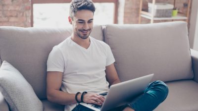 Young attractive smiling guy is browsing at his laptop, sitting at home on the cozy beige sofa at home, wearing casual outfit