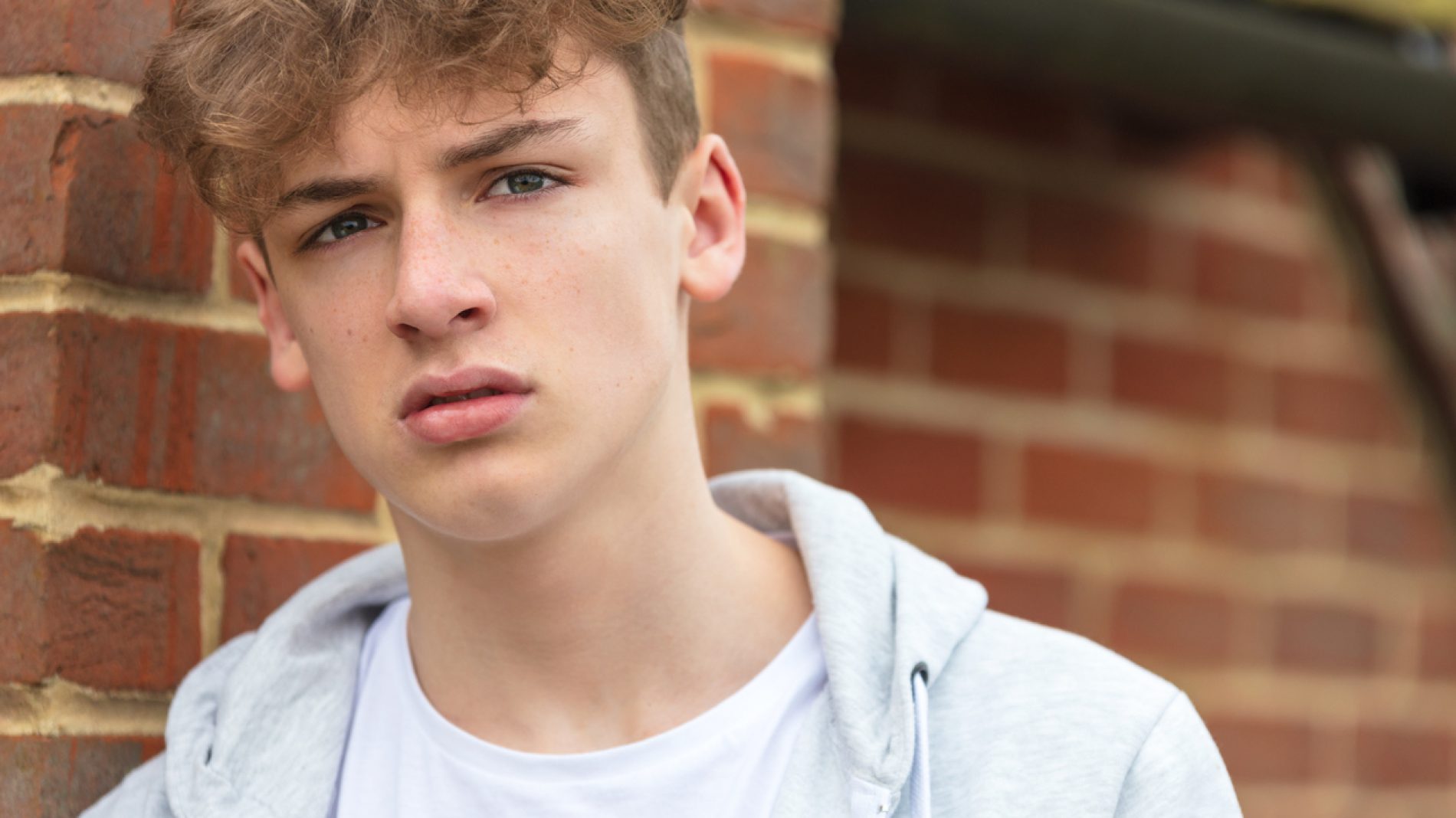 Serious male boy teenager outside leaning on brick wall wearing a gray hoody