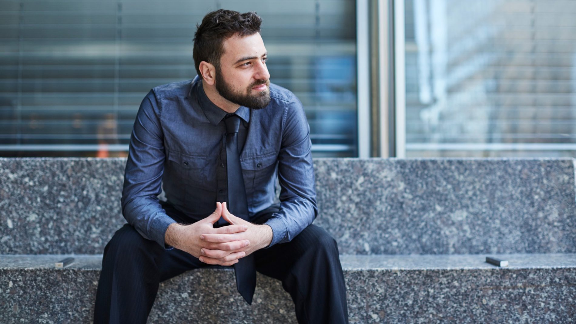 Young bearded Brazilian man leans forward confidently on a bench in the Financial District in New York City financial stress losing job