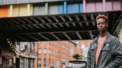 masculine person of colour standing in front of a bridge painted in pride colours. They are wearing a trans pride badge on their black denim jacket. trans ireland