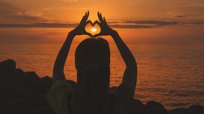 a person is backlit against a sunset. They shape their hands into a heart which surrounds the sun. heartfulness meditation
