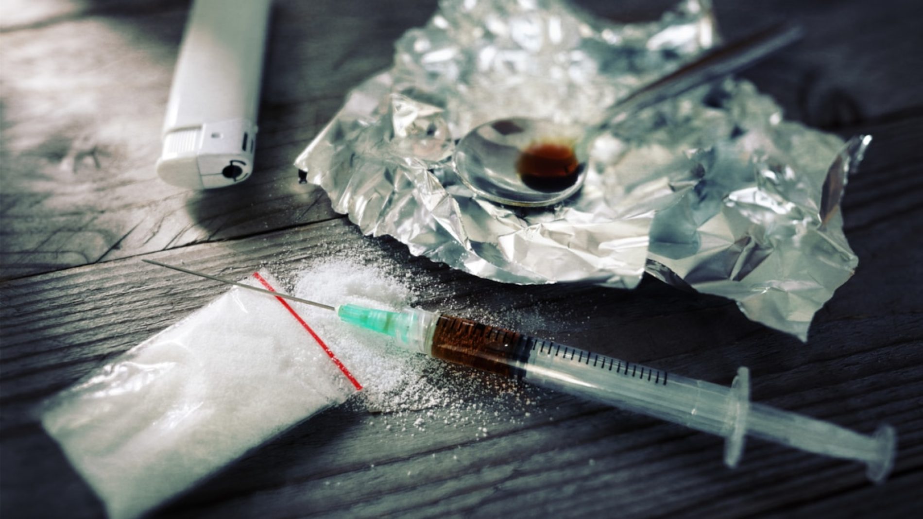Heroin needle full of brown liquid sitting on top of a baggie of white powder, with a white plastic lighter and a crumpled but flattened piece of foil with a teaspoon half-full of brown liquid sitting on top.