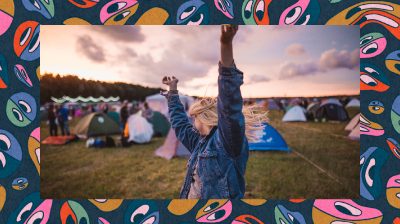 A feminine presenting person raising their arms in the air in front of tents at a music festival