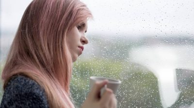 A young person looks out a window holding a cup of coffee