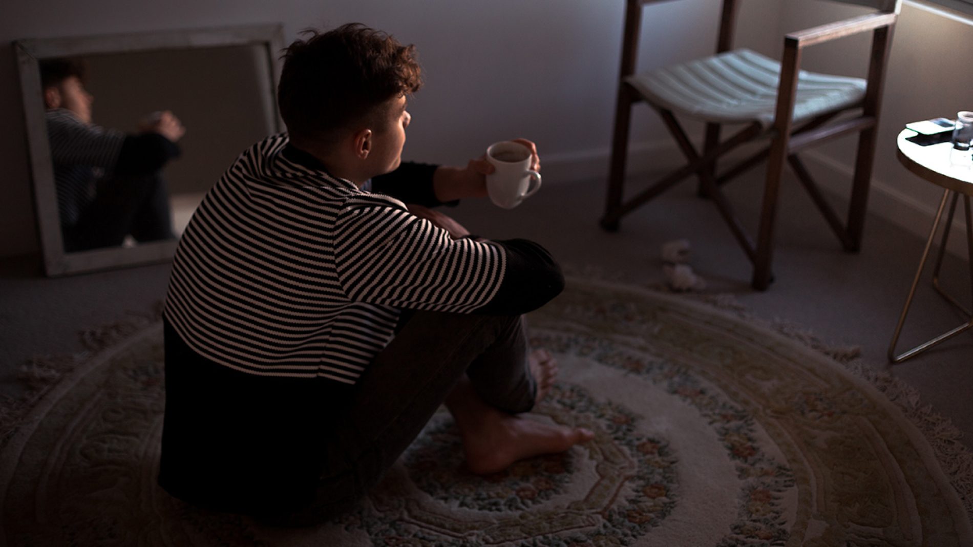 a masculine presenting person sits on a circular rug with their legs crossed holding a cup of tea mindfulness sixth year