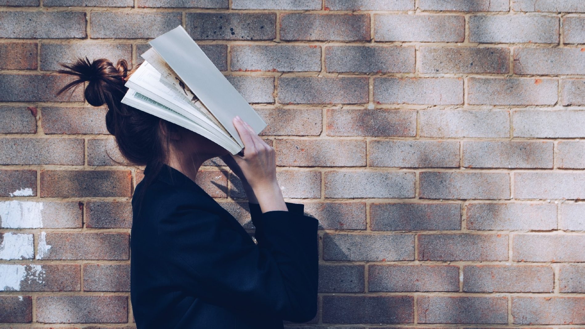 Leaving cert School Girl with Book in front of natural rustic red brick background holding book up to her face.